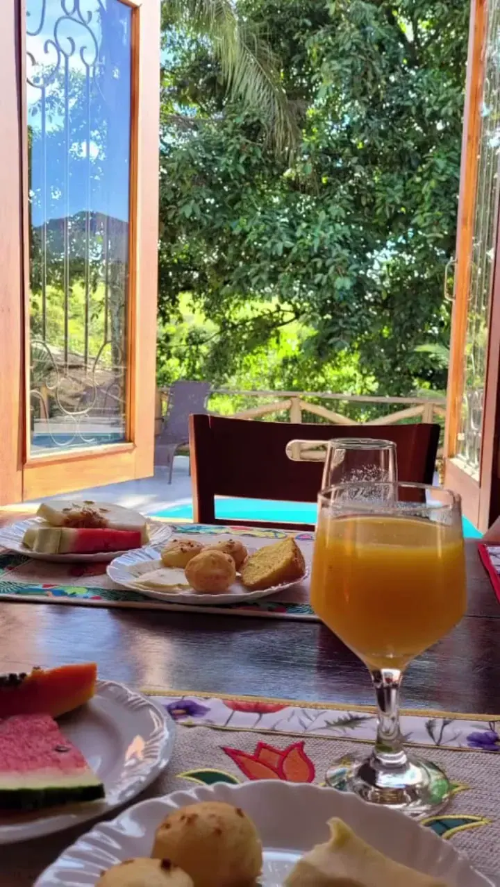 Mesa de café da manhã com suco, frutas e quitandas diante da piscina e do jardim