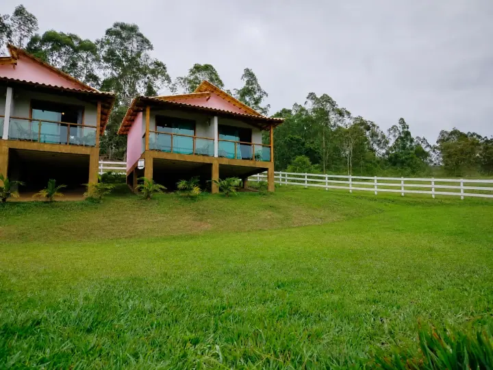 Dois chalés rosa com varandas de vidro diante de um gramado aberto e cerca branca